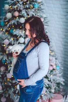 A pregnant girl in a denim jumpsuit with a teddy bear stands near the Christmas tree in the studio