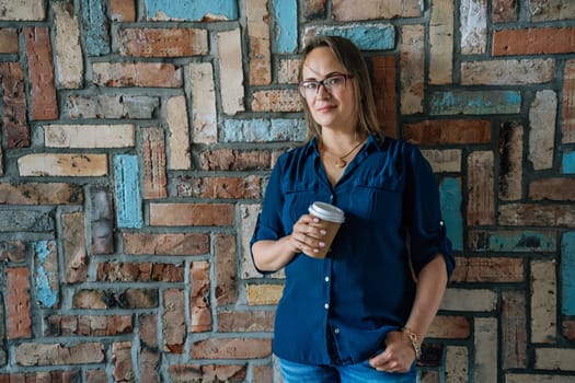 Woman holding a coffee cup, standing against a colorful brick wall. Casual attire, relaxed pose. Concept of leisure and comfort.