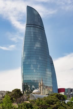 The iconic Flame Towers in Baku, Azerbaijan, rise majestically against a blue sky as seen from Martyrs Lane, resembling flames. Trees in the foreground enhance the cityscape with a natural element