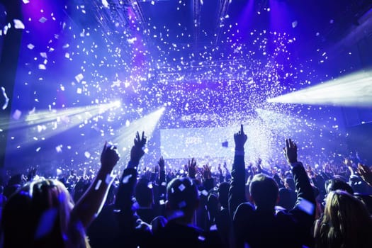 A lively crowd celebrates at a concert hands raised in excitement while confetti falls under vibrant lights filling the venue.