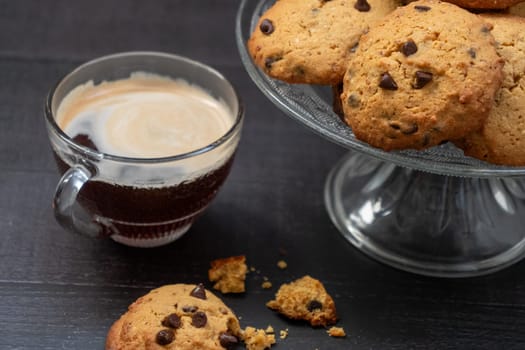 A glass of coffee and a plate of cookies. The cookies are chocolate chip and the plate is on a glass stand