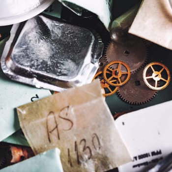 Assorted watch components lie scattered on the workbench, including metal gears, small metal packaging, and labeled paper. Each item reflects the intricate detail of watch repair work