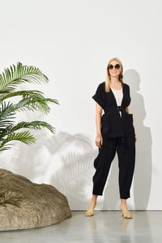 Stylish woman in a black pants and vest paired with white blouse and beige heeled sandals posing in a studio with tropical plant and a rock