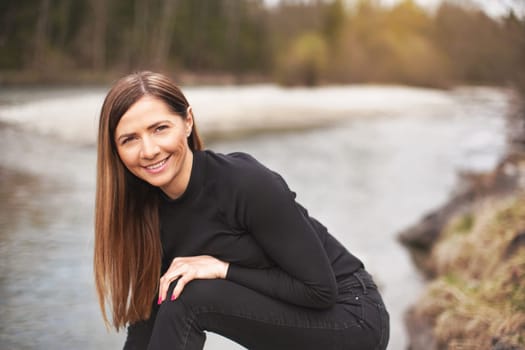 Portrait of young woman wearing black jeans and top, leaning, her long hair falling down, smiling. Blurred river in background