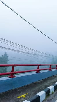A picturesque scene of a foggy landscape viewed from a bridge. Red railings and power lines create a sense of mystery and tranquility in this ethereal setting.