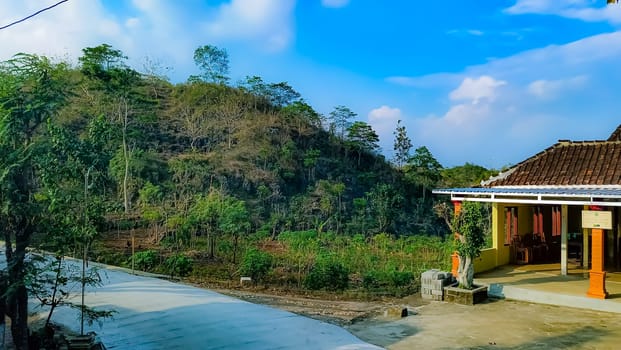 A serene countryside house surrounded by lush vegetation and rolling hills under a bright blue sky, evoking tranquility and natural beauty.