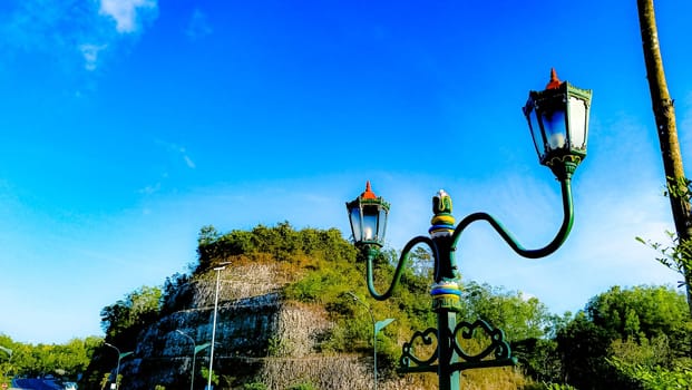 Elegant green street lamps with intricate designs stand in front of a vivid blue sky and lush hill. The scene exudes a blend of nature and historical architecture.