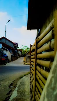 A peaceful village street scene featuring a rustic bamboo fence. The empty road curves gently under a bright blue sky. An ideal setting for themes of simplicity and rural life.