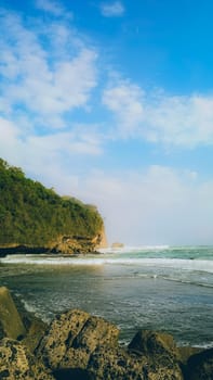 A rocky shoreline with a large rock formation in the background. The water is calm and the sky is blue