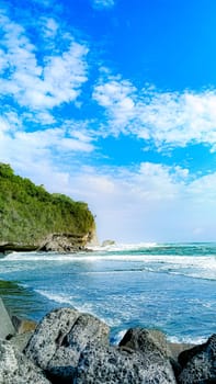 A rocky shoreline with a large rock formation in the background. The water is calm and the sky is blue