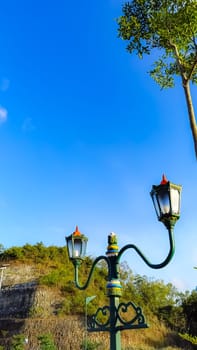 Elegant green street lamps with intricate designs stand in front of a vivid blue sky and lush hill. The scene exudes a blend of nature and historical architecture.