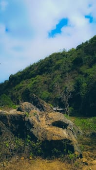 A scenic view of a rocky landscape backed by a lush, green hillside under a partially cloudy sky. The tranquil setting evokes peace and connection with nature.