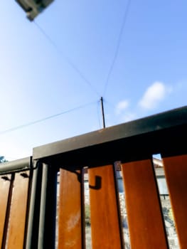 Blurred image showing a wooden fence with metal elements in the foreground. Power lines and a clear blue sky are visible in the background, creating a peaceful and mundane urban scene.