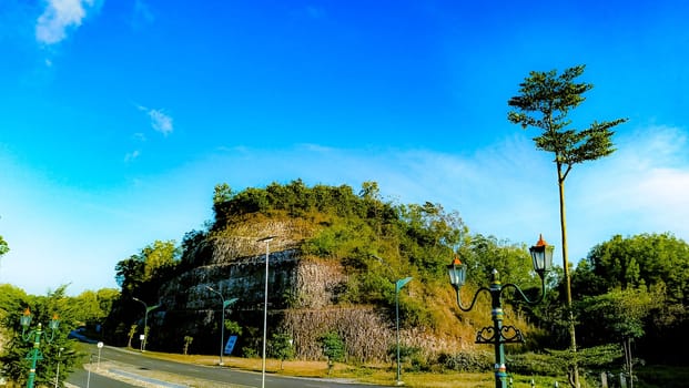 Elegant green street lamps with intricate designs stand in front of a vivid blue sky and lush hill. The scene exudes a blend of nature and historical architecture.