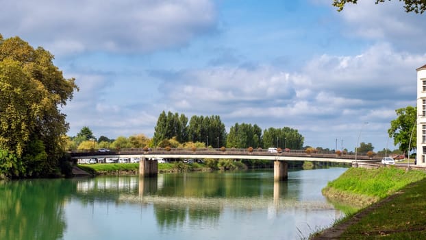 Peyrehorade, France, the bridge spans a river with a green reflection on the water. The bridge is surrounded by trees and there are cars and a truck on the bridge