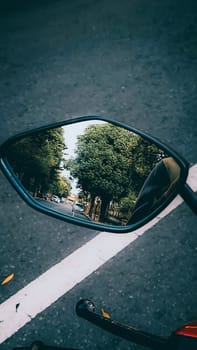 A vivid reflection of a lush tree-lined road is captured in a motorcycle mirror, suggesting travel and adventure. The mirror's angle offers a unique perspective of the picturesque journey.