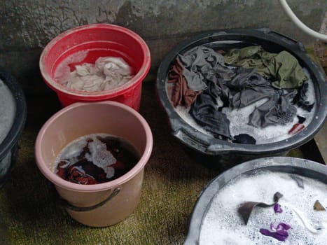 Various clothes soaking in soapy water inside three different colored buckets, depicting a traditional hand washing scene. Vibrant hues and textures convey a simple yet effective laundry method.