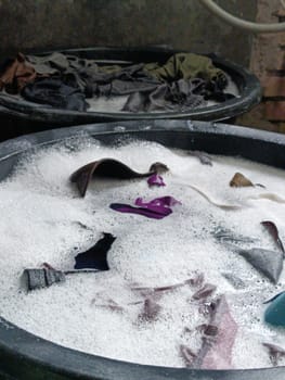 Various clothes soaking in soapy water inside three different colored buckets, depicting a traditional hand washing scene. Vibrant hues and textures convey a simple yet effective laundry method.