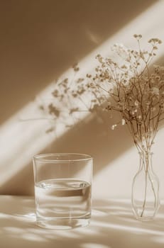 A glass filled with clear water stands next to a slender vase holding soft flowers, highlighted by gentle sunlight patterns.