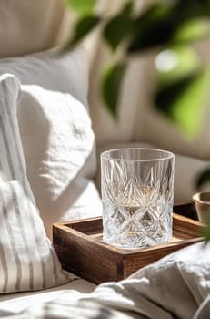 A crystal glass sits on a wooden tray beside soft, textured cushions, illuminated by warm sunlight in a tranquil space.