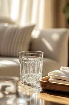 A crystal-clear glass filled with water is placed on a wooden tray next to a cozy couch in a brightly lit living room.