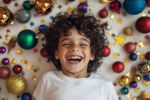 A cheerful young boy smiles widely while relaxing on a surface surrounded by vibrant holiday decorations and ornaments.