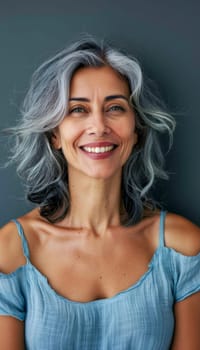 Close-up portrait of smiling middle-aged woman with wavy gray hair wearing light blue top against dark background