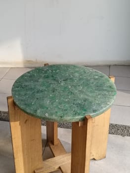A close-up of a round green marble-topped chair with wooden legs placed on a tiled floor in a bright room.