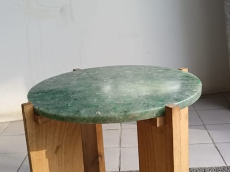 A close-up of a round green marble-topped chair with wooden legs placed on a tiled floor in a bright room.