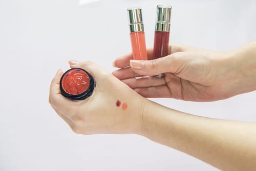 Close-up of hands holding two lip gloss tubes and a jar, showing color swatches applied on the skin for testing.