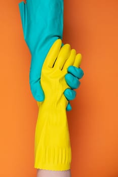 Two hands wearing green and yellow rubber gloves hold each other against a vibrant orange background, symbolizing teamwork and support.