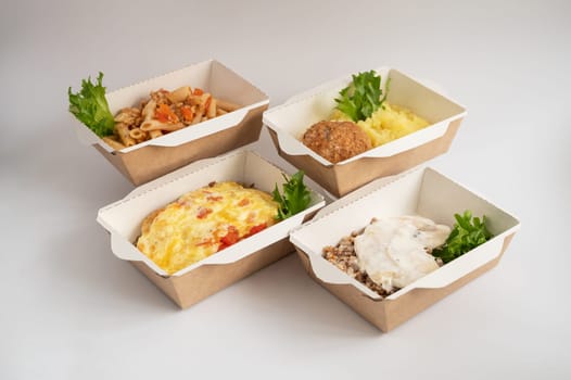Four takeout meals served in biodegradable containers, featuring pasta, casserole, mashed potatoes with meatball, and buckwheat with chicken, garnished with greens.