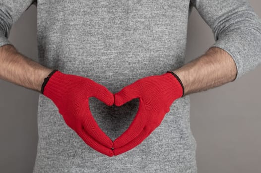 Close-up of hands wearing red gloves creating a heart shape against a gray sweater, symbolizing love, and care.
