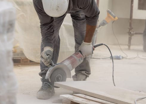 Craftsman working marble with circular saw piece against scaffoldings. Men in dusty grey work clothes and white construction helmet during construction process