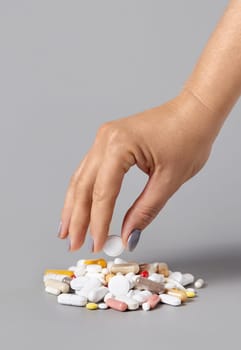 Woman hand put a pillon a pile of medical pills, tablets and capsules closeup against grey background. Assorted pharmaceutical products