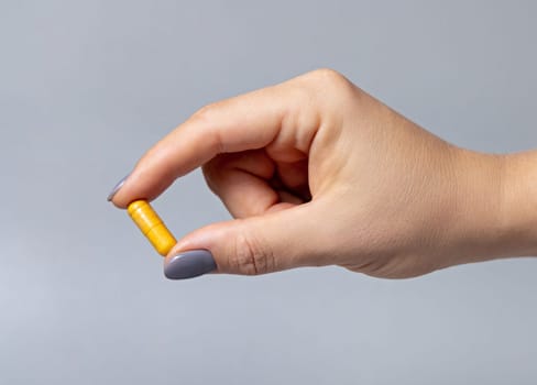 Female hand holding a yellow capsule with fingers closeup against grey background. Healthcare, medical and pharmaceutical concept.