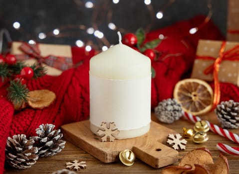 Pillar candle with blank label near red knitted sweater and Christmas decor against lights and wrapped presents, copy space. Warm winter family atmospheric mock up, Holiday time