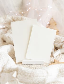 Vertical blank paper cards near white knitted sweater, wooden Christmas decor and lights, copy space. Winter atmospheric mock up, family greeting in rustic or scandinavian style