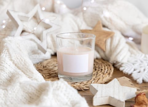Glass candle with blank label near white knitted sweater, wooden Christmas decor and lights, copy space. Winter atmospheric mock up in rustic or scandinavian style