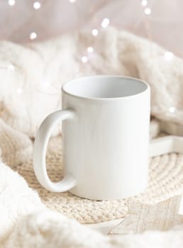 White blank coffee mug near white knitted sweater, wooden Christmas decor and lights, copy space. Winter atmospheric mock up in rustic or scandinavian style