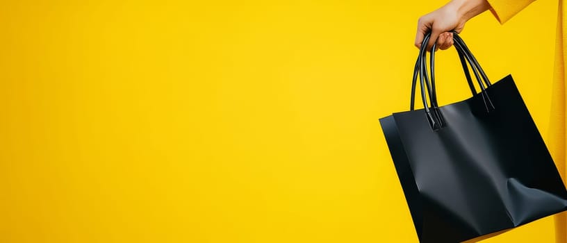 Close-up of a hand holding a black shopping bag against a bright yellow background, symbolizing retail therapy and consumer behavior with copy space.