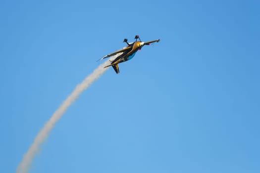 Golden stunt aerobatic plane inverted flying performing stunts in blue sky with smoke trail. Concept of speed, difficulty, precision, risk