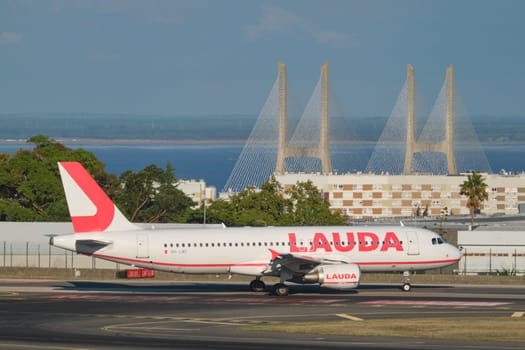 Lisbon, Portugal - September 2, 2023: Lauda Europe Airbus A320-214 passenger plane taxi on runway in Humberto Delgado Airport in Lisbon with Vasco da Gamma bridge in background