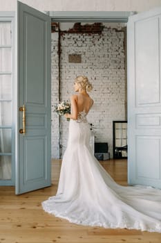 A beautiful bride in an elegant gown holds vibrant flowers, standing by open doors leading to a joyful celebration