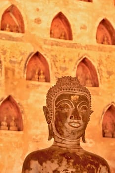 Head of a metal buddha statue, sculpture with a golden, gilded mouth in front of a wall filled with many miniature buddha figurines in the cloister wall of Wat Si Saket, Vientiane, Laos 2023