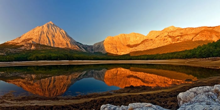 Scenic view of a mountain range reflected in a calm lake during sunrise, with golden hues illuminating the peaks.