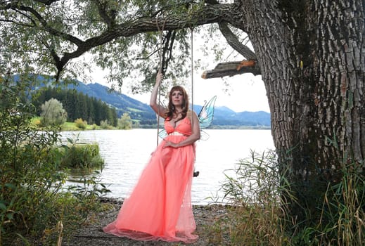 A beautiful elf with a crown and wings sits on a swing by the lake