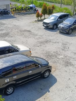 An aerial view of several cars parked in an outdoor lot. The setting is a rural area with visible greenery and distant buildings under a sunny sky.