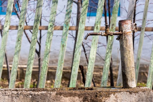 Old iron column. Water is pouring from a street column in front of old green picketed wooden fence.