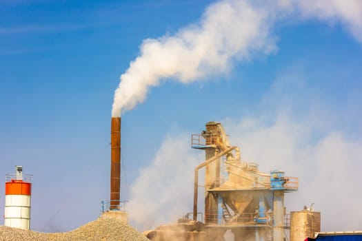 white steam rising up from pipe of small asphalt factory at day time with piles of gravel in a foreground.